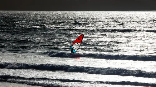 This landscape photograph captures a wind surfer navigating the sea as evening light reflects strongly off the water's surface. It is taken in the summer and shows the wind surfer engaged in surfing atop small waves with the sun low on the horizon, casting a silvery sheen across the ocean. The bright colors of the surf sail contrast against the darker sea, enhancing the visual impact of the wind surfer as the central subject.